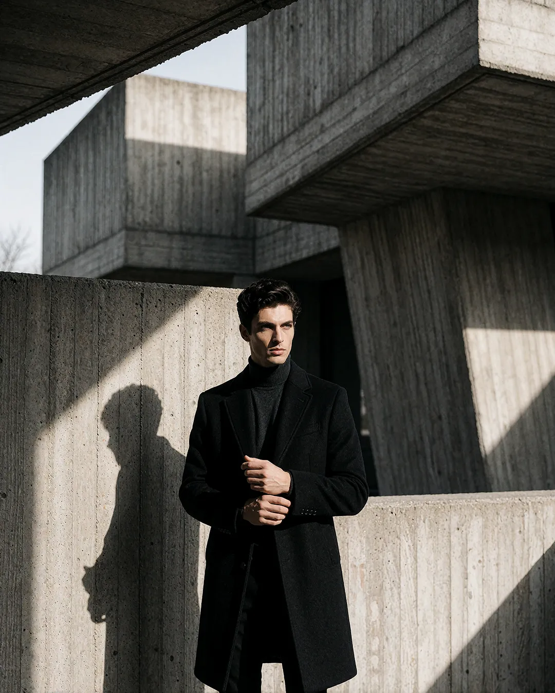 Alessandro Conti, KI Männer Model, im schwarzen Mantel vor brutalistischer Betonarchitektur