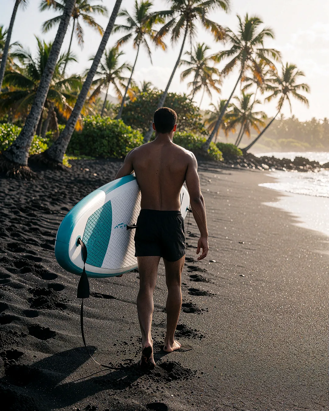 Baptiste Marchois trägt ein Stand-up-Paddel-Board über die Schulter am Strand mit Palmen.