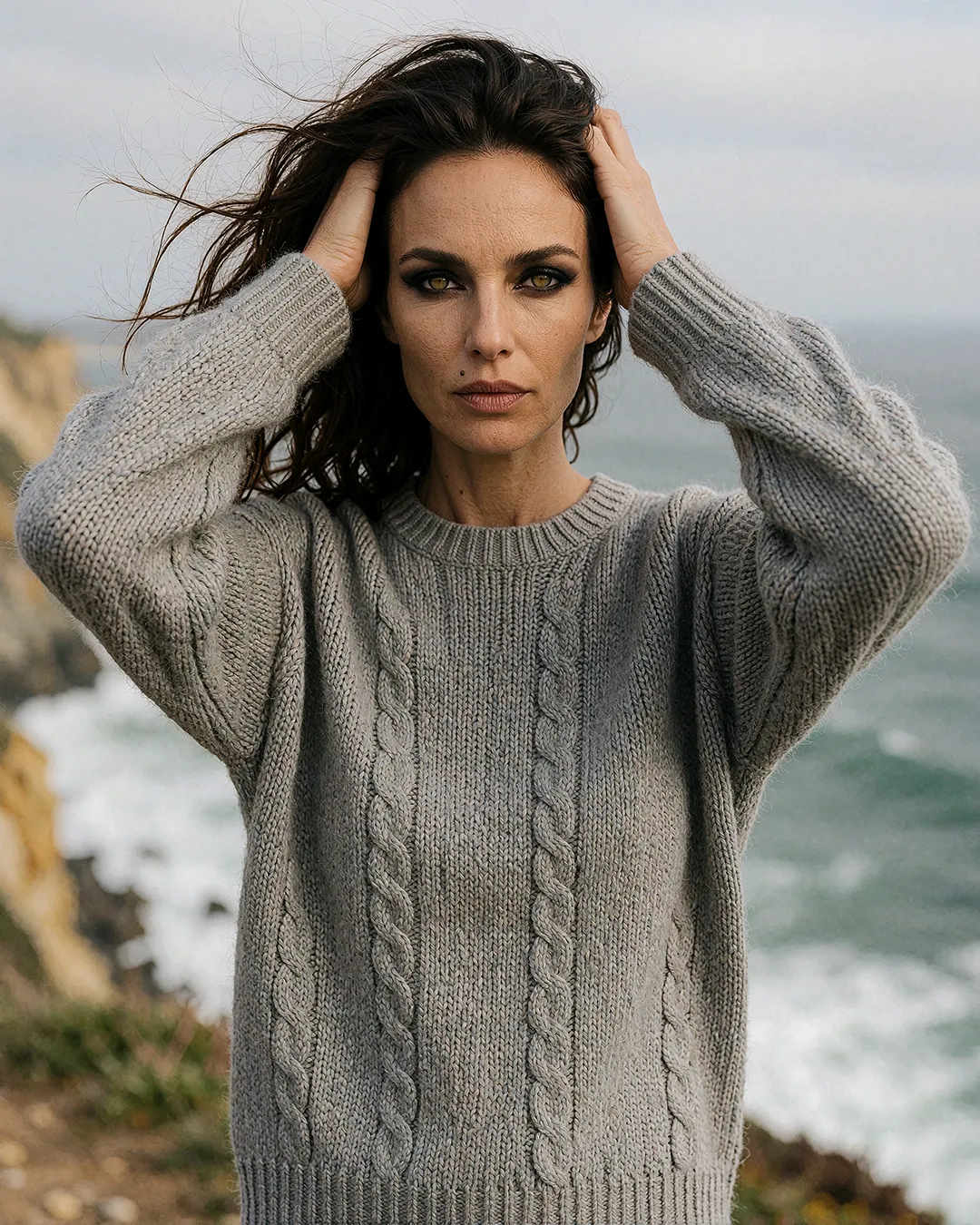 Vivienne Castellane trägt grauen Zopfstrickpullover mit windzerzaustem Haar an der Steilküste.