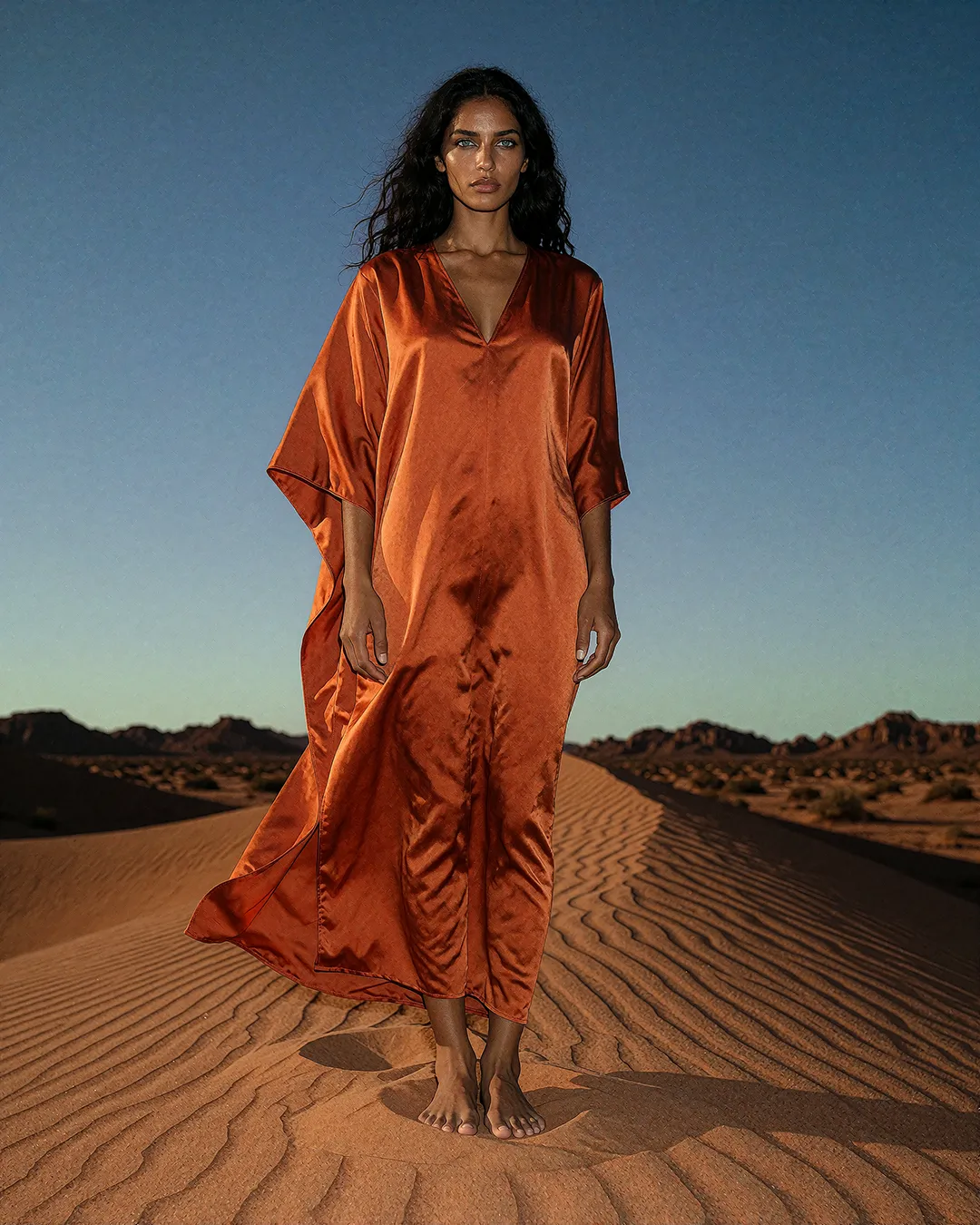 Samira Farrouk steht barfuß in kupferfarbenem Satin-Kaftan auf einer Wüstendüne bei Dämmerung.