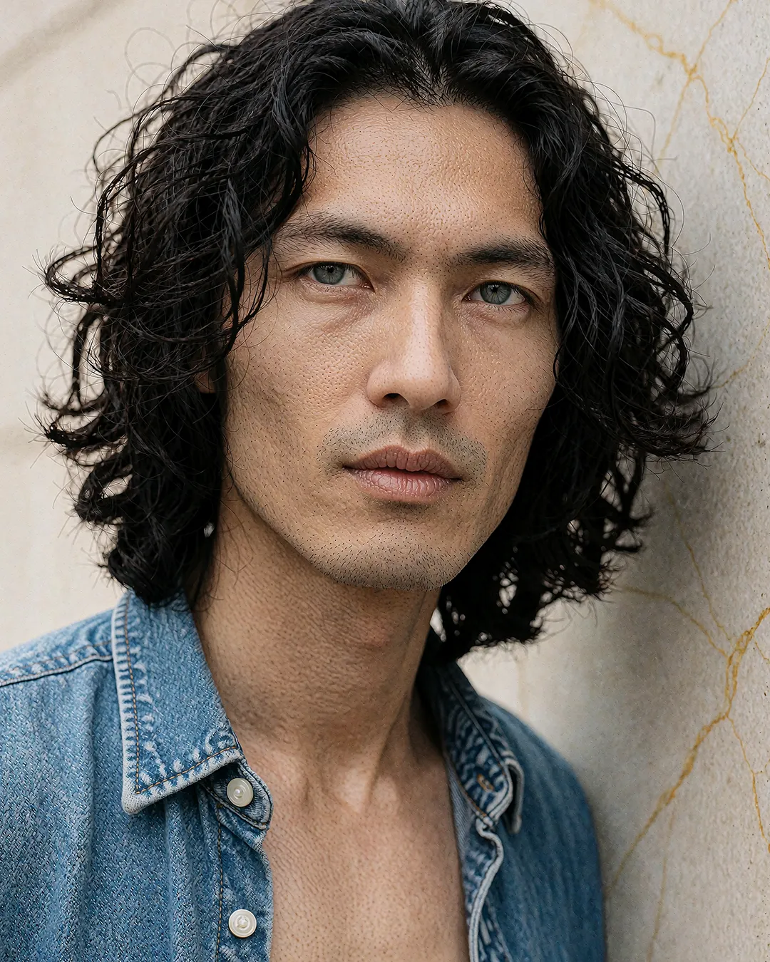 Kenji Osato im offenen Jeanshemd vor rissiger Steinwand, Nahporträt