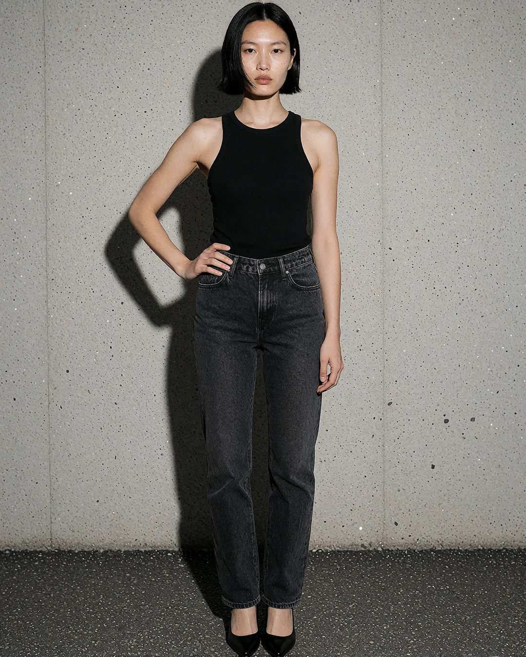 Nanami Onodera lehnt lässig in schwarzem Top und Jeans an einer Betonsäule.