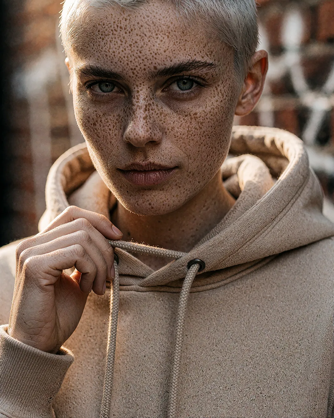 Robin Aschenberg im Nahporträt mit Sommersprossen, sandfarbener Hoodie vor Backsteinmauer.