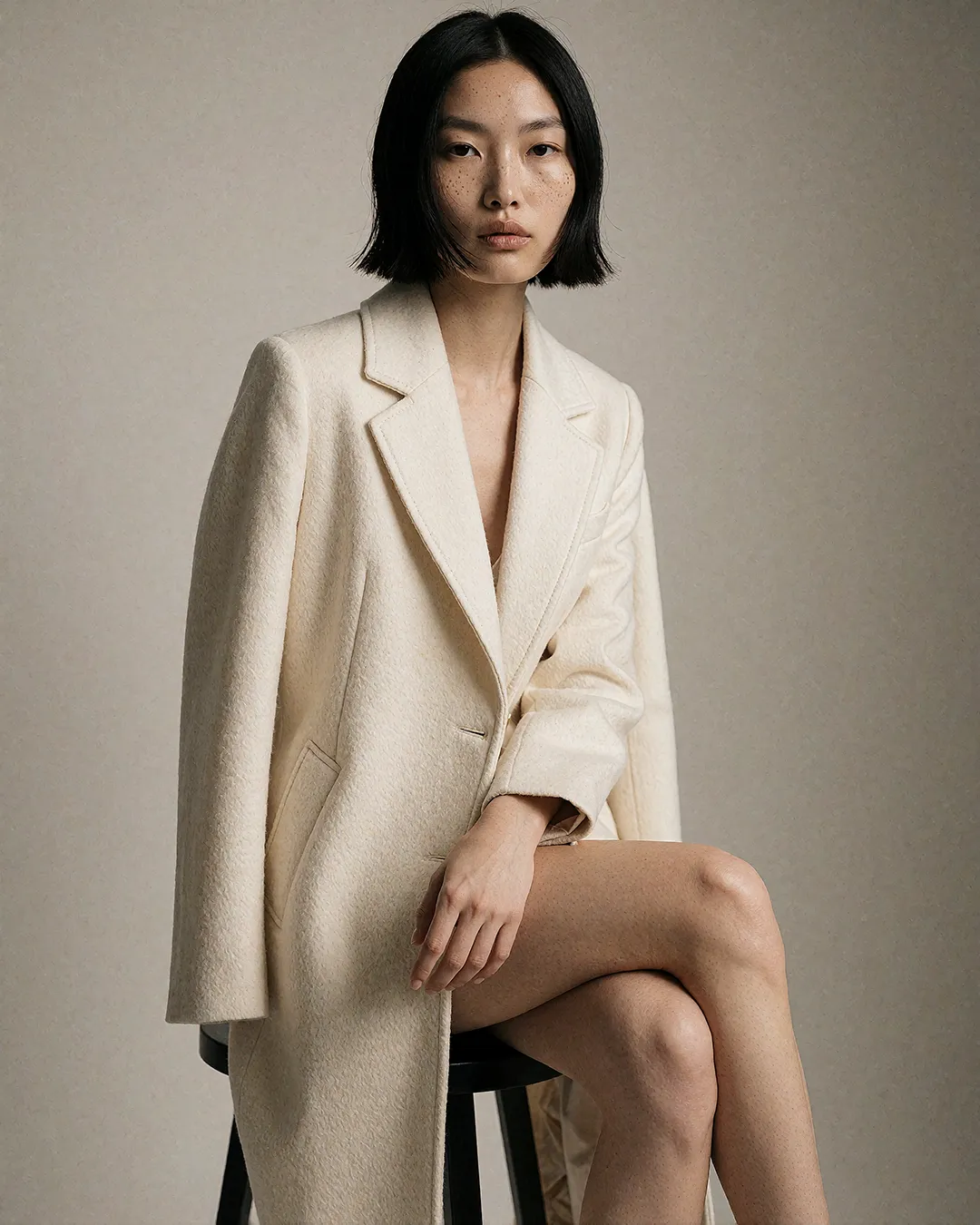 Nanami Onodera sitzt im cremefarbenen Oversize-Mantel elegant auf einem Hocker im Studio.