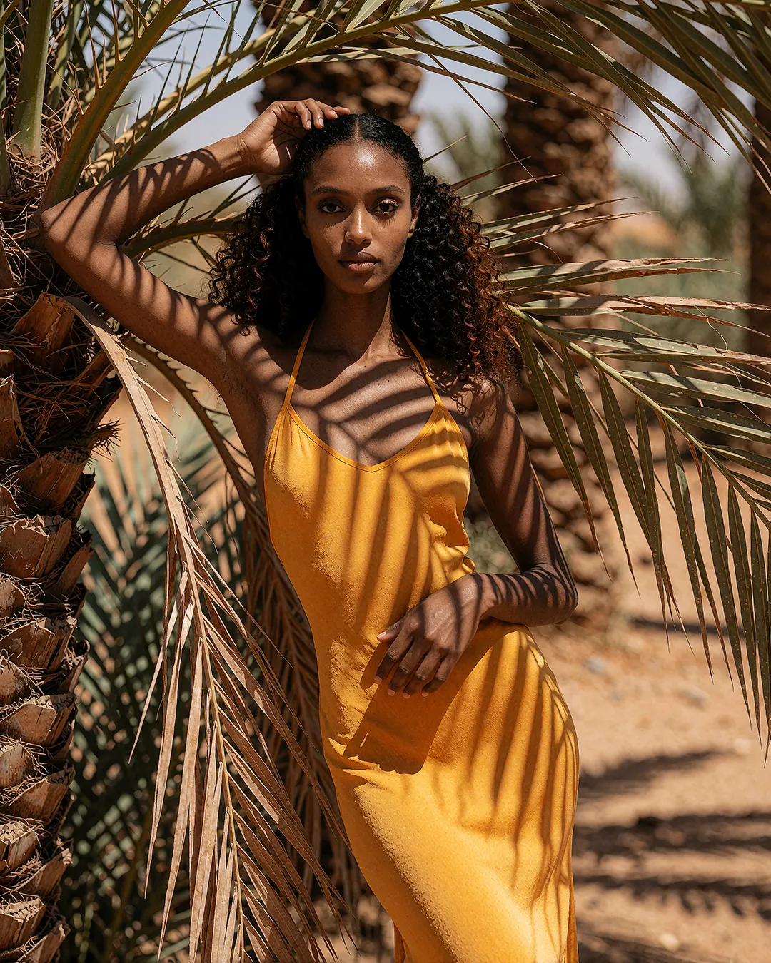 Zuri Osei trägt ein senfgelbes Seidenkleid im Schattenspiel einer Palmenoase.