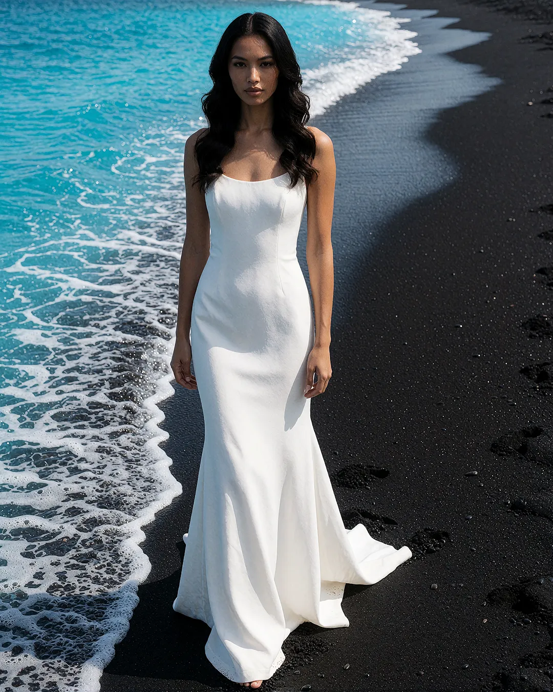 Mika Thornbury trägt ein weißes Trägerkleid am schwarzen Vulkanstrand mit türkisfarbenem Meer.