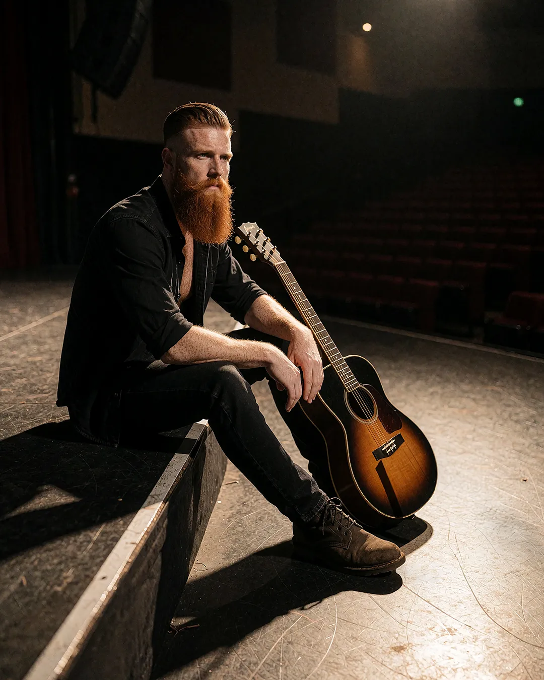 Ronan Flaherty sitzt mit Akustikgitarre nachdenklich auf der leeren Bühne eines Theaters.