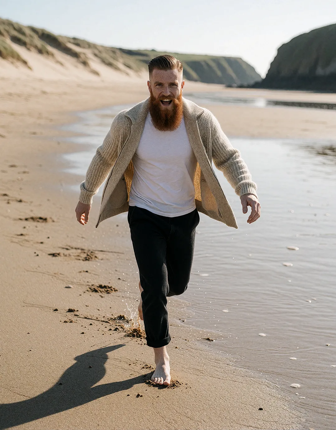 Ronan Flaherty läuft lachend barfuß im offenen Cardigan durch die Brandung am Sandstrand.